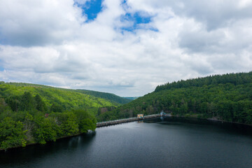 Fototapeta premium A tranquil reservoir is bordered by a concrete dam and surrounded by dense, vibrant forested hills beneath a sky filled with dramatic clouds at Lac de Chaumecon.