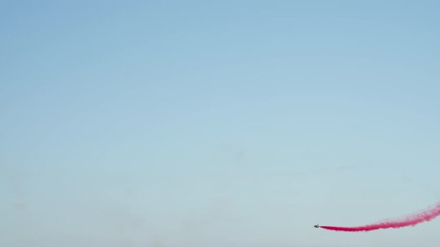 An aerobatic jet performs a maneuver, leaving a red smoke trail. It crosses paths with another jet trailing blue smoke in a spectacular precision airshow display against a clear blue sky.