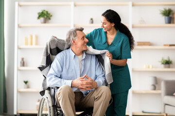 Obraz premium A woman with long dark hair smiles while helping a man with gray hair in a wheelchair. They are in a well-lit room with plants and shelves. The caregiver drapes a blanket over the man.
