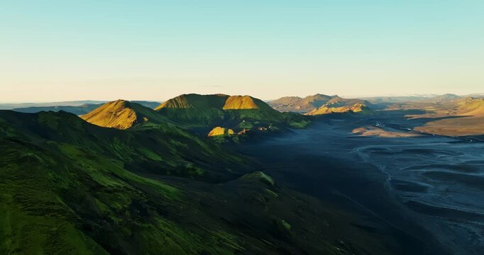 Golden-hour drone glide over a mossy caldera and black sand valley in Iceland highlands, with braided river patterns and calm, epic wilderness under a clear sky.