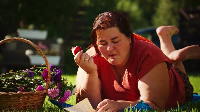 A woman enjoying a sunny day in the garden, lying on a blanket, surrounded by flowers and reading a book while holding a fresh fruit in her hand.