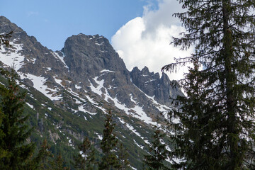 Tatra Mountains landscape in Poland featuring rocky peaks, snow patches, and lush green coniferous trees under a partly cloudy sky