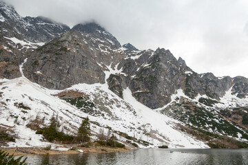 Morskie Oko lake surrounded by snow-capped Tatra Mountains in Poland, showcasing rocky cliffs and a serene water surface reflecting the cloudy sky
