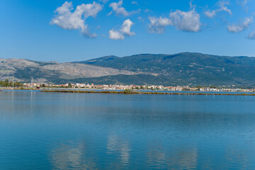 Fototapeta premium A calm salt lagoon in western Greece reflects the blue sky and scattered clouds, with the town of Missolonghi and distant mountain slopes forming a scenic backdrop. The tranquil water and geometric