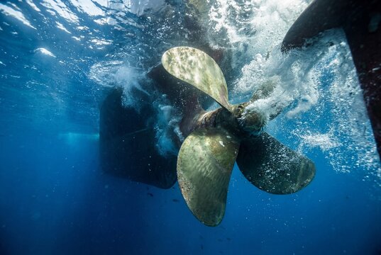 Underwater view of large bronze ship propeller with bubbles and sunlight in deep blue ocean water
