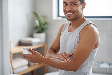 Cheerful young man applying white lotion to his forearm for skincare in a bright domestic bathroom
