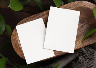 Vertical cards on brown wooden tray near green leaves and grey fabric top view, boho mockup