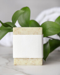 Mockup of beige solid soap bar with paper label and green leaves near white bath towels, personal care