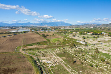 Naklejka premium Aerial perspective of the archaeological site of Paestum, Italy, featuring expansive stone foundations and ruins set beside cultivated fields. The scene is framed by distant mountains under a clear