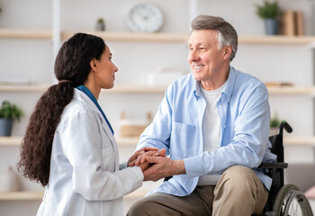 Obraz premium A medical professional talks to a senior man using a wheelchair. They are in a bright therapy room with soft decorations. The atmosphere shows support and care.