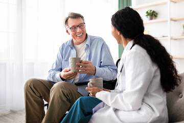 Obraz premium An older man with gray hair and glasses sits in a wheelchair smiling at a woman who is talking to him. They are holding mugs in a sunny room filled with plants and light.