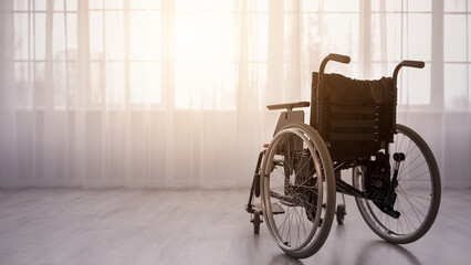 A wheelchair stands alone in a bright room filled with natural light coming through large windows. The atmosphere suggests an absence, inviting thoughts of the person who usually occupies it. © Prostock-studio