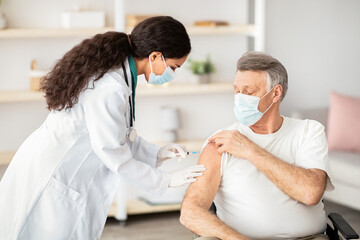 Obraz premium A health worker assists an older man with gray hair in receiving a vaccine. Both are wearing masks, and the setting is a well-lit clinic. The health worker is focused on the task.
