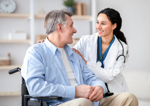 A healthcare professional smiles and talks to an older man sitting in a wheelchair. They are in a well-lit room with simple decor. The mood is friendly and supportive, focusing on the conversation.