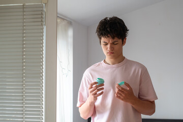 Young man holding skin cream and frowning, teenager's daily acne care routine, problematic young skin concept