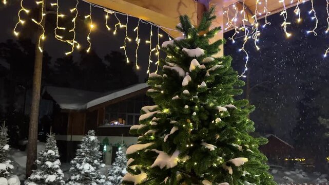 Snowy Christmas tree with glowing string lights at night. Winter holiday decoration for festive atmosphere. Cozy outdoor scene. New year eve celebration background. Cozy home village.