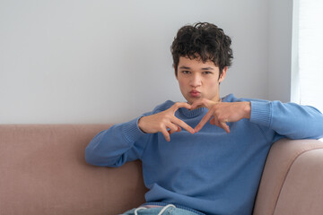 A cute young man is showing a love sign, forming a heart with his fingers, making a kiss with his lips, wearing a stylish blue sweater, sitting on a pink sofa. Body language concept