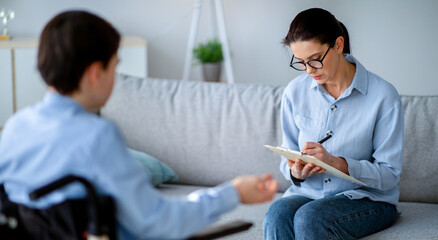 Obraz premium Woman with glasses sits on a couch writing on a clipboard while listening to a man using a wheelchair. They are in a modern living room setting with light colors.