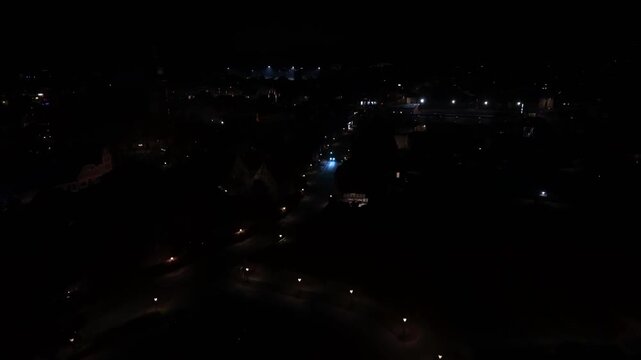 Aerial night view of residential area with moving cars and glowing headlights, showing traffic assert motion through dark streets. Light patterns across American neighborhood. Forward wide shot.