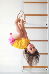 Active little girl hanging upside down on gymnastic rings at home, smiling child in bright clothes enjoying indoor physical activity, childhood development, play healthy lifestyle concept © Evgeniia Primavera