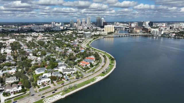 Drone view of Hyde Park in Tampa, Florida, showing coastal waterfront homes, curved shoreline roads, calm bay waters and downtown skyline under bright daytime sky in USA. Wide shot.