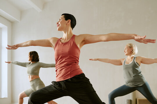 Three Caucasian women practicing yoga in studio, standing in warrior pose with arms extended, including two middle aged women and one young adult woman focusing on balance and flexibility