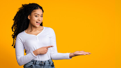 Young African American woman stands against a bright yellow background, smiling and pointing to her left. She wears a long-sleeve white shirt and denim jeans. © Prostock-studio
