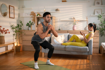 Obraz premium Man doing squat exercise while woman relaxes on sofa 