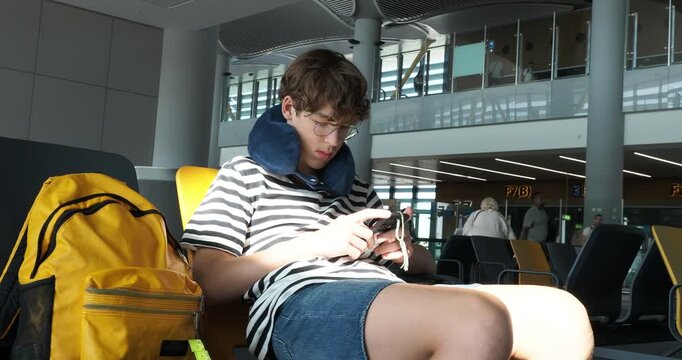 A young man with a neck pillow sits in an airport terminal, engrossed in his smartphone while waiting for his flight. A relatable scene of modern travel, boredom, and digital connectivity.
