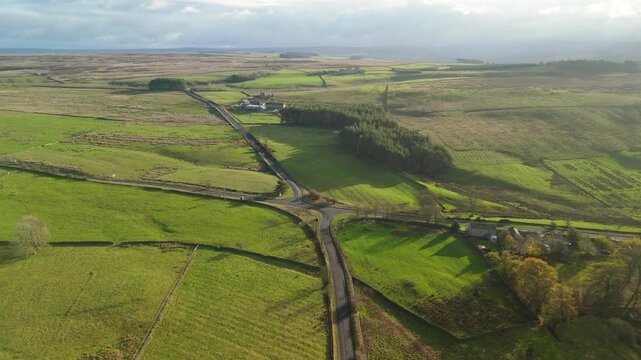 A drone captures a view of green fields and winding roads in Northumberland near Hexham. The landscape features trees and a farmhouse in the distance under a clear sky