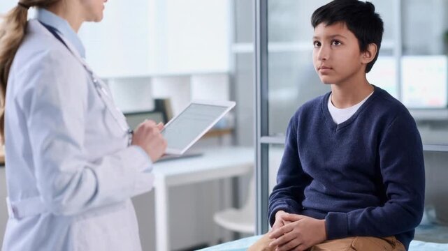 Teenage boy sitting on examination table nodding answering pediatricians questions while doctor using digital tablet at doctors office