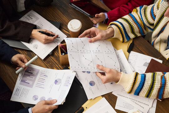 Group of young adult and middle aged Caucasian and Black hands analyzing business charts and graphs at table, holding printed documents with data, discussing financial strategy, coffee cup visible
