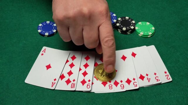 Hand places a gold coin bitcoin on a row of playing cards featuring diamonds on a green felt table surrounded by colorful poker chips