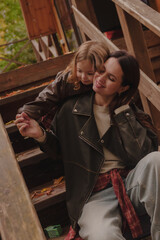 Mother and daughter hugging on the steps of a wooden staircase. Cheerful girls in the country in the fall. A little girl hugs her mother from behind. Vertical photo.