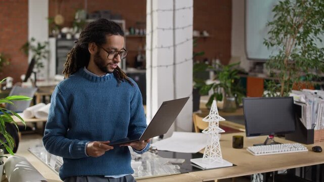 Medium portrait of Biracial male engineer standing in bright modern office holding laptop with architectural and engineering layouts on table in background