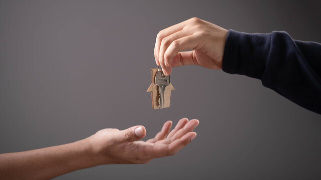 Hand giving house keys with wooden home keychain to another hand, symbolizing real estate deal, property handover, home ownership, buying house, mortgage approval, trust, and housing business concept.