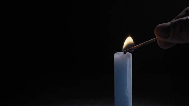 Hand lights a single candle with a match against a black background. Concept of hope, remembrance, memorial, spirituality, and quiet contemplation.