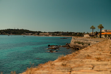 Fototapeta premium Beautiful Mediterranean coastline with turquoise sea, stone promenade and palm trees on a sunny summer day. Scenic seaside landscape, travel destination concept, calm and relaxing atmosphere.
