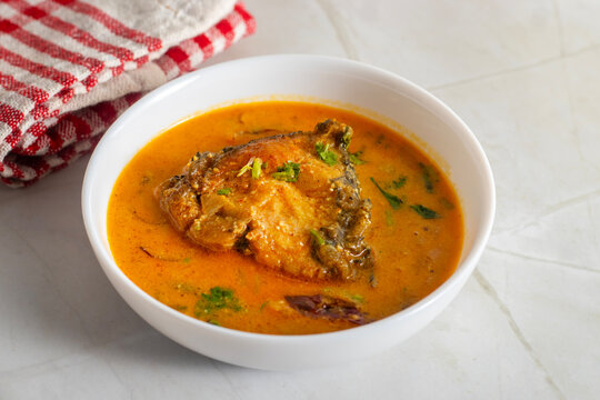White bowl holds Indian katla/catla fish curry Bengali style with golden gravy, tender fish pieces, onion slices, and herbs, styled on marble surface with red cloth background.
