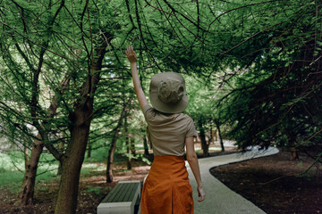 Obraz premium Woman in forest wearing a hat and orange skirt reaching up to tree branches on a winding path by a bench, exploring nature during a quiet summer walk in green woodland alone.