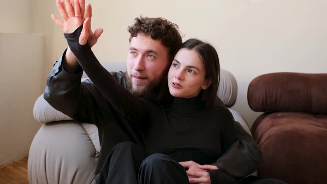 Young happy couple embracing and cuddling together on a sofa at home, with the man holding the woman's hand while she looks outside, sharing an intimate and loving moment in their living room
