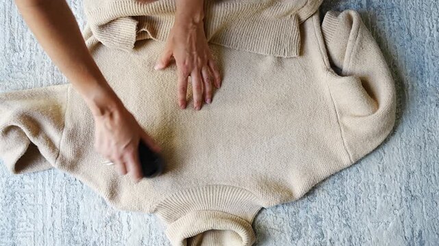 Top view of woman removing lint from sweater with fabric shaver on floor at home. Clothing care process, fabric maintenance and practical preparation before wearing.