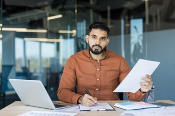 Obraz premium Young professional man sitting at an office desk, reviewing documents and writing on a clipboard, managing important business tasks with a laptop nearby