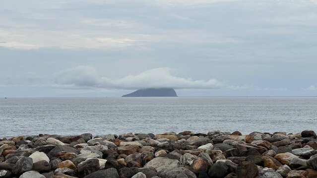 A tranquil view of Guishan Island in Taiwan, A rocky shoreline foreground with colorful stones, leading to a calm ocean and a distant island partially covered by clouds under a gray sky.