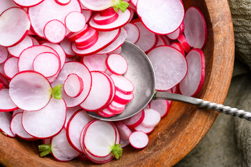 Sliced red radishes in wooden bowl with metal spoon on textured fabric background. Fresh salad ingredients for healthy diet and rustic food photography