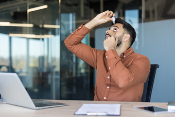 Young man applying eye drops to his irritated eyes at his office desk, finding relief from dryness...