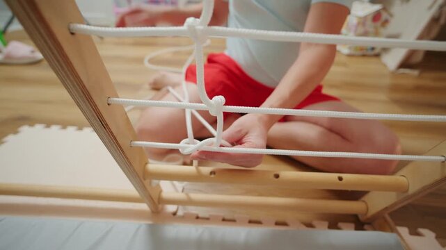 Person ties knots on a wooden ladder during an indoor activity in a bright room with colorful toys in the background