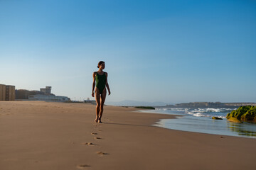 Athletic woman in a green swimsuit walking barefoot along a tranquil beach in morning light, leaving a trail of footprints in the sand, with ocean waves and soft blue sky behind her.