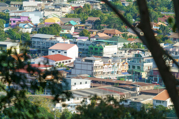 High-angle view of a vibrant Southeast Asian town nestled among trees. Colorful residential and commercial buildings create a dense urban landscape under bright daylight. Professional travel photo.