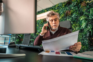 Mature man communicating on phone, managing paperwork, and analyzing data in an office with a green wall
